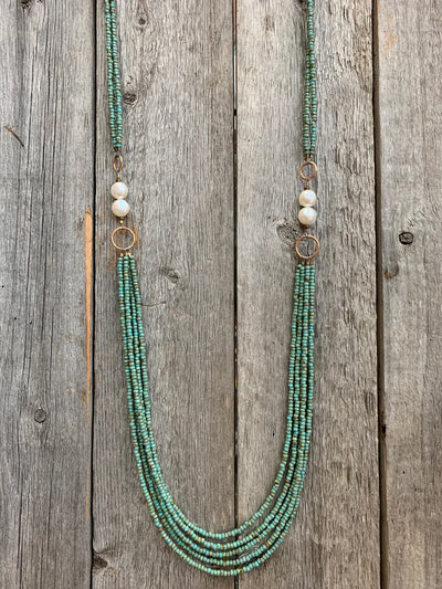J.Forks Designs Necklace hanging on wooden backdrop. This necklace is a five-strand 32" turquoise seed bead necklace with freshwater pearl sides and solid bronze hook clasp.