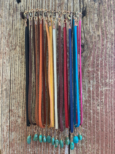 J.Forks Designs multi-color leather necklaces hanging on a wire in front of a wooden backdrop. Each necklace is a 15" Leather Necklace with with Hammered Bronze Ring and Kingman Turquoise Drop. The necklaces are in the following order from left to right, 