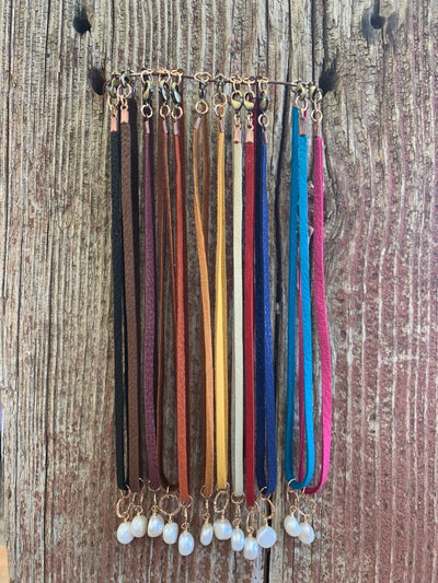 J.Forks Designs necklaces hanging on wooden backdrop. The necklaces are 15" Colored Leather with Bronze Hammered Ring and Freshwater Pearl Drop. The necklaces are hanging in the following order: Black, Chocolate, Wine, Saddle Tan, Burnt Orange, Cognac, Mu