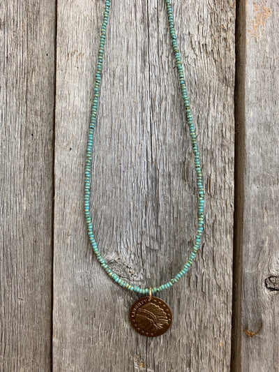 J.Forks Designs necklace hanging on wooden backdrop. This is a 15" turquoise seed bead necklace with bronze Indian coin pendant and bronze lobster claw clasp.