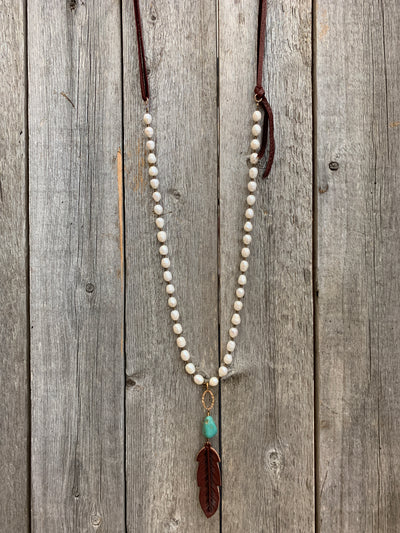 J.Forks Designs necklace hanging on wooden backdrop. This is a 32" Freshwater Pearl and wine leather back necklace with Kingman Turquoise and hand-tooled wine leather feather.