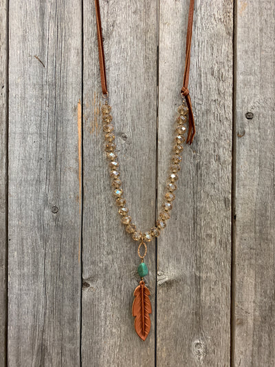 J.Forks Designs necklace hanging on wooden backdrop. This is a 30" Golden Austrian Crystal and chestnut leather back necklace with Kingman Turquoise and hand-tooled chestnut leather feather. 