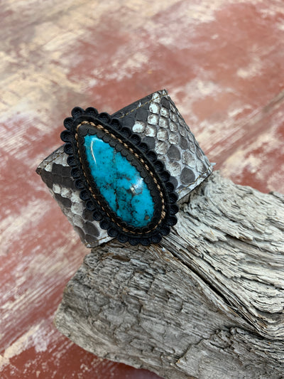 J.Forks Designs 1 1/2" wide Natural Python Cuff with a 45-carat Bisbee Turquoise stone that is hand set in black leather. The bracelet is placed on a wooden stump and showcases the turquoise stone.