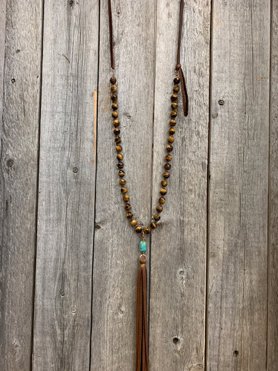 J.Forks Designs necklace hanging on wooden backdrop. This a 32" Tigers Eye necklace with a Kingman Turquoise drop, chocolate leather tassel, and chocolate leather back.