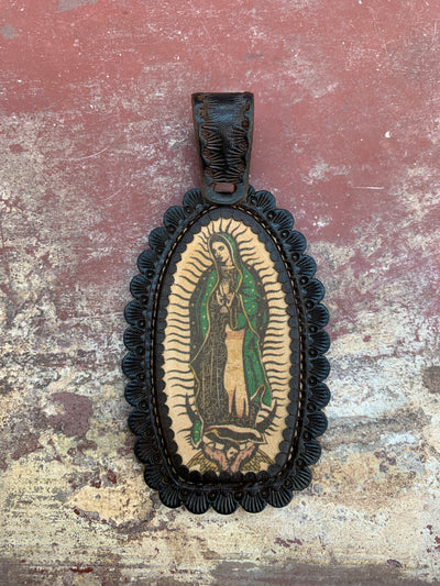 This J.Forks Designs pendant is laying on concrete backdrop. This pendant has an Our Lady of Guadalupe stone that is hand set in chocolate leather. This pendant is 5" long from top of bail and 2.5" wide. 