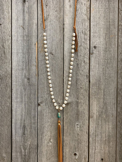 This is an image of the J.Forks Designs 32" White Freshwater Pearl Necklace with Kingman Turquoise and Cognac Leather Tassel hanging on a vintage wood background. This photograph was taken at the J.Forks Designs studio on Main Street in Boerne, Texas. 