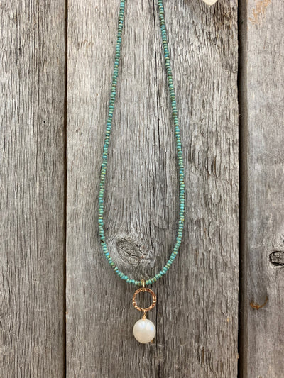 J.Forks Designs necklace hanging on a wooden backdrop. This is a 16" turquoise seed beads necklace with a hammered bronze ring and freshwater pearl drop.