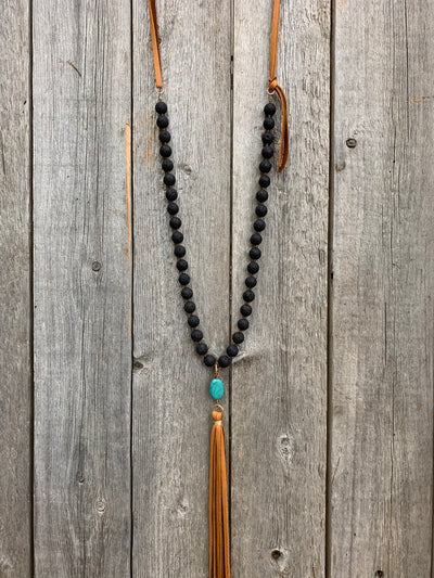 J.Forks Designs necklace hanging on wooden backdrop. This is a 32" lava rock and cognac leather back necklace with Turquoise and leather tassel drop.