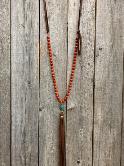 J.Forks Designs Necklace hanging on a wooden backdrop. This necklace is 28" with small red sponge coral and chocolate leather back, turquoise, and leather tassel drop