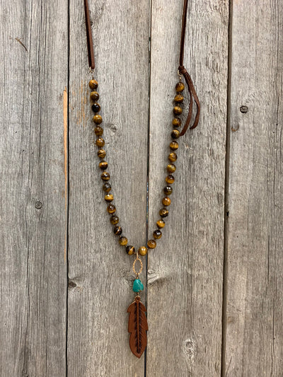 J.Forks Designs necklace hanging on wooden backdrop. This is a 28" tigers eye and leather back necklace with turquoise and hand tooled chocolate leather feather drop.